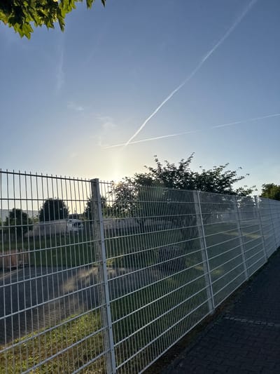 The image depicts a wire fence along a paved walkway, with greenery and trees visible behind the fence. The sky is blue with two distinct contrails from aircraft. The sun is low in the sky, casting a warm light and creating some lens flare. In the background, there are some buildings and vehicles. The scene appears to be an outdoor area, possibly a park or an industrial zone, in Bad Säckingen, Germany.