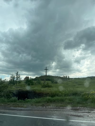 An exterior, eye-level, medium shot captures a cross monument situated atop a grassy hill under a cloudy sky. To the left of the cross, a stone structure, possibly a church or ruins, is visible among trees. The foreground features a dark, overgrown ditch and a wet asphalt road with a white line. The overall scene is overcast and appears to be in Artashavan, Armenia.