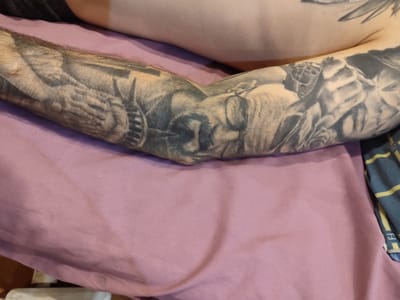 The media file is a close-up, eye-level shot of a person's arm covered in extensive black and grey tattoos. The arm rests on a pale pink fabric surface, likely a bed or couch. The tattoos depict a complex, narrative scene, featuring a portrait of a person wearing glasses, possibly an older man, with intricate shading and detail. Surrounding the portrait are various elements including what appear to be gears or machinery, floral motifs like roses, and possibly other figures or symbols, creating a dense and visually rich composition. The lighting appears to be neutral, with no strong shadows, suggesting an indoor setting during daylight hours. The focus is entirely on the tattooed arm and the artwork itself, with the background blurred. There is no visible text in the image. The context of Lisakovsk, Kazakhstan, does not offer any specific visual cues within the image itself.