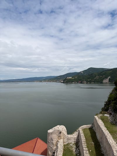 The image shows a wide river bordered by hills and a fortified structure on a cliff overlooking the water. The river is a dull green color, reflecting the cloudy sky. In the distance, a town or village is visible along the riverbank, nestled at the foot of the hills. The hills are covered in dense green vegetation.

In the foreground, a section of a stone fortification, likely part of a castle, is visible. The stones are rough and weathered, with patches of green grass growing in between them and along the top of the walls. A red-tiled roof of a building is also visible in the lower left corner. The overall impression is of a historical site with a dramatic natural backdrop. The location is identified as Golubac Fortress, in the Golubac Municipality of Serbia.