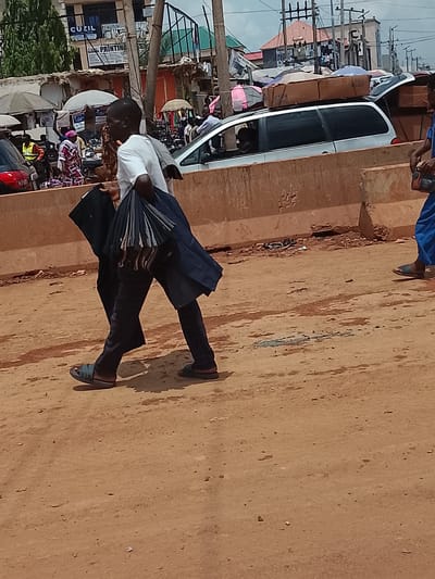 The image shows a man walking on a dusty, brown-colored ground surface in an urban area. He is wearing a white shirt, dark-colored pants, and sandals, and he is carrying a dark-colored garment or object in his left hand and what appears to be a bundle of umbrellas or similar items in his right. Behind him are indications of a bustling street scene, including people, vehicles, and vendor stalls with umbrellas. A light-colored minivan with boxes or items stacked on its roof is partially visible, as is a section of a concrete barrier. The background includes buildings, utility poles, and various signs. The location is identified as Katambe, Nigeria.