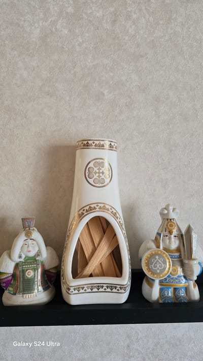 A neutral-toned, textured wallpaper serves as the backdrop for a shelf displaying three decorative items. On the left is a porcelain figurine of a person in traditional attire, featuring a white fur-lined hat and ornate green, gold, and purple clothing. Next to it is a tall, white, vase-like object with gold trim and a circular emblem on its front. The opening of this vase contains a bundle of light-colored, thin wooden sticks. To the right of the vase is another porcelain figurine, this one depicting a warrior-like figure with a white and gold headdress, holding a sword in its right hand and a shield adorned with a gold emblem in its left. The warrior is dressed in blue and gold clothing. The shelf itself is black. The bottom left corner of the image displays the text "Galaxy S24 Ultra". The scene is indoors, suggesting it is a domestic setting. There is no indication of the specific location in Khatassy, Russia.