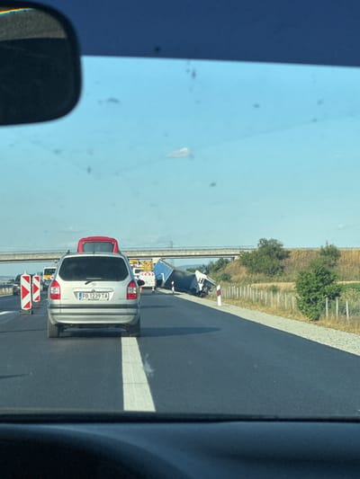The image captures a highway scene in Bozduganovo, Bulgaria. The perspective is from inside a vehicle, looking out and slightly to the right. The road ahead shows a traffic slowdown, likely due to an accident or roadwork, indicated by a white truck with hazard lights and construction barriers. In the foreground, a silver car with license plate "PB 1239 TA" is visible, its rear facing the camera. Behind it, a red vehicle is partially obscured. The road is paved and gray, with a textured shoulder and a guardrail separating the lanes from the dry, grassy embankment beyond. A blue sky with scattered small dark spots, possibly dirt or debris on the camera lens, dominates the upper portion of the image. Trees and vegetation are present on the embankment. The lighting suggests it is daytime, likely late morning or afternoon, given the bright sunlight casting shadows. The overall impression is of a routine commute interrupted by unexpected traffic delays on a sunny day in a rural highway setting.
