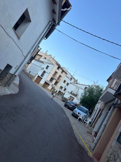 The image shows a street scene in Alpuente, Spain. The street is paved and slopes upwards. On the left side of the street, there is a white building with a small, narrow window. On the right side, there are several buildings with white facades and tiled roofs. Some of the buildings have balconies and windows. There are two cars parked on the right side of the street, a grey Audi and a silver car. In front of the silver car, there are two white plastic chairs. A young woman wearing a white dress is walking up the street, carrying a potted plant. The sky is clear and blue. The overall impression is of a quiet, sun-drenched village street.