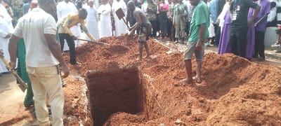 The image captures an outdoor scene in Umu Okoro Uku, Nigeria, depicting a group of people gathered around a freshly dug grave, likely during a burial or funeral service. The ground is characterized by prominent reddish-brown soil, piled high around a deep, rectangular excavation.

In the foreground and center, several men are actively engaged with the grave. A man in a white polo shirt and light-colored trousers stands with his back to the camera, holding a shovel. To his right, another man in a yellow patterned long-sleeved shirt and dark pants is also using a shovel to move earth. A shirtless man in patterned shorts is actively digging with a shovel, standing directly at the edge of the deep pit. Further to the right, a man in a green t-shirt and light shorts stands observing near a mound of soil.

A significant crowd of onlookers surrounds the grave area, observing the proceedings. Among them are several individuals dressed in white religious robes, suggesting the presence of clergy. Other attendees are dressed in various civilian clothes, some appearing formal, and a few individuals are seen in dark suits or military-style uniforms. The scene is brightly lit by natural sunlight, indicating it is daytime. The overall atmosphere appears solemn, consistent with a funeral gathering. No distinct or readable text is visible on any subject in the image.