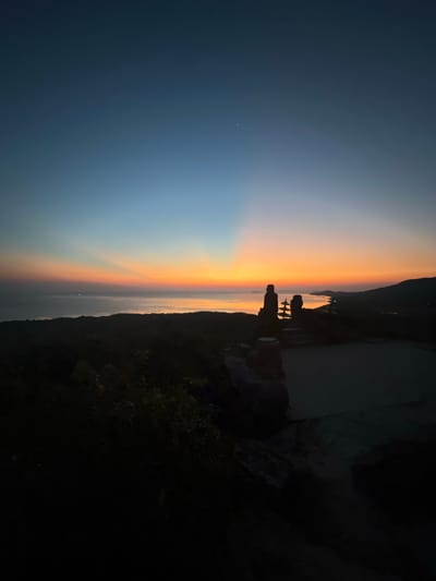 The image depicts a silhouetted landscape at sunset. The foreground features dark, shadowy foliage and a stone structure that resembles a small statue or monument. Beyond this, a body of water, likely the sea, is visible, with distant lights suggesting a coastal settlement or scattered boats. The sky transitions from a deep blue at the top to vibrant hues of orange, yellow, and pink near the horizon, indicative of a sunset. Rays of light fan out from the setting sun. The overall scene evokes a sense of tranquility and natural beauty. The location is identified as Karimunjawa, Indonesia. No text is visible in the image.