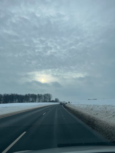 The scene depicts a winter landscape on a rural highway in Shelepino, Russia, viewed from inside a vehicle. The sky is overcast with dense, grey clouds, through which a diffused, bright light source, consistent with the sun, is visible on the left side. A multi-lane asphalt road, appearing wet or slushy, extends into the distance, curving slightly to the right. White lane markings are discernible. On the left side of the road, a line of bare deciduous trees stands against a snow-covered field. On the right, a dark-stained snowbank borders the road, giving way to an expansive, snow-covered field in the midground. Several vehicles, including what appear to be trucks, are visible in the distance on the highway. The immediate foreground shows a portion of a dark vehicle dashboard or windshield frame. No people are visible.