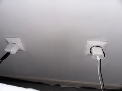 The image shows a white ceiling with two electrical outlets. Each outlet has a white power adapter plugged in. The left outlet has a white adapter with a black cable connected, and a small blue light is visible on the adapter. The right outlet has a white adapter with a white cable connected. The scene appears to be indoors, likely a room in Shalkia, Kazakhstan, given the prompt. The lighting is dim, casting shadows on the wall below the outlets.