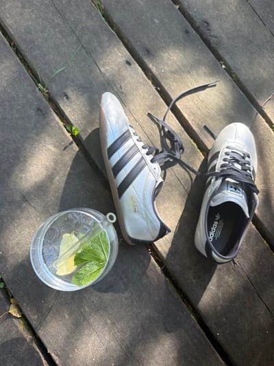 Two silver Adidas sneakers, one with black laces trailing, are placed on a weathered wooden deck. Next to the shoes, a clear glass contains ice, lime slices, and mint leaves, with a glass straw inserted. The scene is outdoors, with sunlight casting shadows across the deck, suggesting a warm day. There are glimpses of greenery and fallen leaves along the edges of the deck, indicating a natural or garden setting. The specific location in Mühlbach - Rio di Pusteria, Italy is not discernible from the visual information provided in the image. There are no people present.