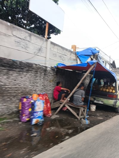 The image captures an outdoor scene in Dayeuhkolot, Indonesia, alongside a paved road. In the midground, a makeshift vendor stall is set against a textured, dark-stained wall, which transitions to a lighter concrete section with graffiti above. The stall comprises wooden poles supporting blue and red tarpaulins that provide shade.
Two men are seated on simple wooden benches within the shaded area. The man on the left wears a red short-sleeved shirt and dark shorts, facing right, holding a small object to his mouth. The man on the right wears a grey striped short-sleeved shirt and dark pants, seated with his back partially to the viewer, facing a light green minivan. The minivan is parked with its rear hatch open, revealing numerous whole coconuts stacked inside. A white bucket is present on the ground near the minivan's rear wheel.
To the left of the stall, several large, colorful plastic bags, including those appearing to be pet food (one labeled "CAT"), are stacked on wet ground containing puddles. A blank white billboard is mounted on a wooden post above the wall on the left side of the frame. Power lines are visible against the cloudy sky in the upper portion of the image. Green foliage from trees is present above the wall on the far left.