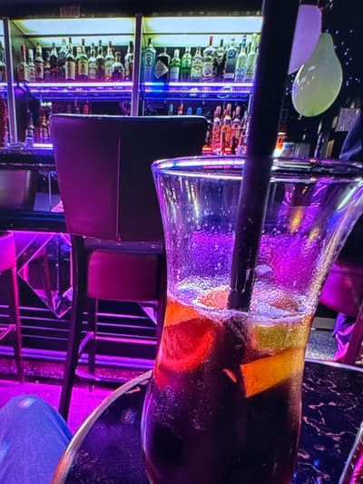 The image depicts a close-up view of a cocktail in a tall glass, with a dark beverage and slices of fruit such as oranges and possibly grapefruit. A black straw is visible in the drink. The setting appears to be a bar, with shelves stocked with numerous bottles of liquor and spirits in the background. The area is illuminated with vibrant purple and blue neon lights, creating a lively atmosphere. In the mid-ground, a dark-colored bar stool with a purple cushion is partially visible. Further in the background, there are balloons, one of which is light green and partially deflated. The image is captured from a slightly elevated angle, focusing on the drink and the immediate surroundings. There is no discernible indication of the location being in Porrentruy, Switzerland.