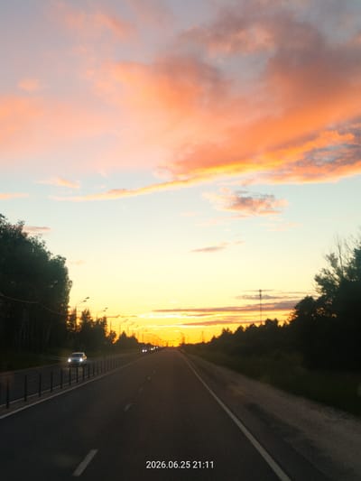 The image captures a scenic view of a road at sunset in Ulyanino, Russia. The sky is ablaze with vibrant hues of orange and pink, transitioning into a soft yellow and then a pale blue as it reaches higher. Fluffy clouds are scattered across the sky, catching the last rays of the sun and creating a dramatic and colorful display.
The road itself is asphalt and appears to be a highway or a major road, with a clear division of lanes marked by white lines. On the left side of the road, a dark line of trees forms a dense barrier, with streetlights intermittently visible among them. On the right, more trees and vegetation line the roadside, with a tall, slender tower-like structure visible in the distance.
Several vehicles are present on the road. A white car with its headlights on is driving in the left lane, approaching the viewer. Further down the road, more vehicles can be seen as distant lights. The overall mood is one of tranquility and beauty, with the sunset creating a peaceful and awe-inspiring atmosphere.
Visible text at the bottom right corner of the image reads "2026.06.25 21:11", indicating the date and time the photograph was taken. This information, combined with the scenic view, suggests the photograph was taken during an evening drive.
