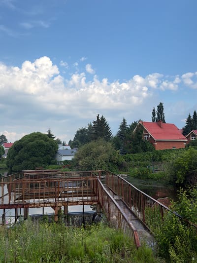 The image depicts a rural landscape in the village of Maloye Sedelnikovo, Russia, on a clear day with scattered clouds. In the foreground, a rusty, weathered pedestrian bridge with metal railings extends over a body of water. Tall green grass and vegetation grow along the banks of the water. In the mid-ground, a large, dense green tree stands prominently. Beyond the tree, several houses are visible, including one with a distinctive red tiled roof and brick facade. The houses are surrounded by lush greenery and trees. The sky above is a vibrant blue with fluffy white clouds.