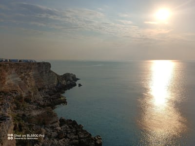 The image depicts a coastal cliff overlooking the Black Sea at Kamen Bryag, Bulgaria. A line of several white camper vans is parked along the edge of the cliff on the left side of the frame. The sea, with its surface rippling under the bright sun, occupies the majority of the image. The sun is high in the sky, casting a strong reflection on the water, creating a shimmering path of light. The cliff face is rocky and textured, with patches of vegetation visible in some areas. The sky above is partially cloudy, with wispy white clouds scattered across a pale blue expanse. The overall scene captures a serene coastal landscape bathed in sunlight.