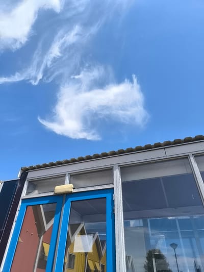 A low-angle, medium shot captures a building's facade against a bright blue sky filled with wispy white clouds. The building appears to be modern, with a corrugated roofline visible at the top. Below the roof, a series of windows form the upper part of the structure. One window frame is a bright blue, with glass panes reflecting a colorful building with a red and yellow pitched roof. To the right of the blue-framed section, a larger window with dark-tinted glass reflects the sky and a streetlamp. The scene is devoid of any people.