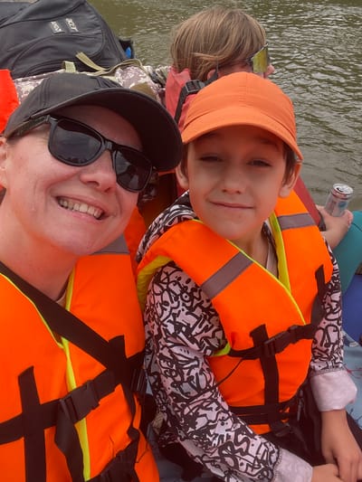 In a medium shot, a group of people are seated on a raft on a body of water, likely in or near Samorodovo, Russia. The foreground is dominated by two individuals wearing bright orange life vests. The person on the left, facing the camera, wears a black baseball cap and dark sunglasses. They have a wide smile, revealing their teeth, and their skin appears slightly flushed. Beside them, on the right, is a child wearing an orange baseball cap and a life vest. The child is looking towards the camera with a gentle smile. Behind them, partially visible, is another individual wearing sunglasses. A black backpack with "AMD" and "acer" logos is visible in the background, along with bags and life vests. The water appears murky and calm. The lighting suggests an outdoor daytime setting.