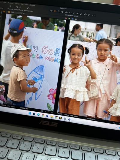 The image is a close-up, taken from a slightly elevated angle, of a laptop screen displaying a digital editing program. The screen shows two photographs side-by-side. The left photograph depicts a young boy wearing a blue and red baseball cap and a light brown t-shirt, holding a blue marker and drawing on a large white surface, possibly a banner or a wall. He is focused on drawing a blue rocket ship. Text in Russian is visible on the surface, partially obscured. To the right of this boy, the top of another child's head is visible.

The right photograph displays three young girls. The two girls in the foreground are facing the camera. The girl on the left has pigtails with orange ribbons and face paint. She is holding up two fingers in a peace sign with her right hand. The girl on the right is also making a peace sign with her right hand and is wearing a light pink checkered outfit. Between them, a small, pink purse with pearl embellishments is visible. A third girl, only partially visible on the far right, is also wearing white clothing with ruffles. The background of both photos suggests an outdoor event with other people milling around, some of whom are blurred.

The laptop itself is a silver ASUS model. The keyboard is visible in the foreground, showing standard QWERTY layout. The top bar of the editing program displays menus and tools, including the program name "Capture One." Image file names like "DSC08667.JPG" and "DSC08848.JPG" are visible at the bottom of the screen, along with camera settings such as "1/500 сек f/4.5 46 mm" and "1/250 сек f/4.5 28 mm".

The location is identified as Novopokrovka, Kyrgyzstan. The scene appears to be a community event or festival, likely involving children and art activities.