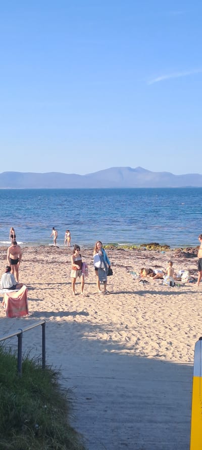 This is a vertical, eye-level shot of a beach scene on a sunny day, looking out towards the sea and a distant mountain range. The sand is light brown and textured with ripples. In the foreground, a metal railing with two vertical posts borders a patch of green grass. The sea is a vibrant blue, with small waves lapping at the shore. Several people are scattered across the beach and in the shallow water.
In the mid-ground, there are two women walking on the sand. The woman on the left is wearing a brown and white patterned top and light-colored shorts, carrying a pink bag. The woman on the right is wearing a blue jacket over dark clothing and carrying a grey towel and a dark bag. To their left, a man in black swim trunks stands on the sand, facing the water. Further to the left, a person in a light-colored shirt and dark shorts sits on a red towel. In the water, several people are visible, some wading and others further out.
In the background, the sea stretches to the horizon, meeting a hazy, purple mountain range under a clear blue sky. The overall impression is a relaxed, summer day at the beach in Kilsallagh, Ireland.