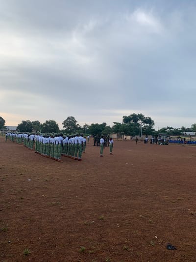The image displays an outdoor scene in Kubi A, Nigeria, under an overcast sky. A large formation of approximately several dozen individuals stands in multiple straight rows across a reddish-brown soil field with sparse green grass. These individuals are uniformly dressed in white short-sleeved shirts, light green trousers, and brown footwear, primarily facing away from the camera. In the mid-ground, to the right of the main formation, several individuals in dark green camouflage uniforms, possibly supervisory personnel, are observed. Additionally, other individuals in similar white and green attire are present. The background features a line of mature green trees, with light-colored buildings partially visible behind them. On the far right, a temporary assembly area includes rows of blue plastic chairs and a green canopy. The scene depicts a formal outdoor assembly or training exercise.