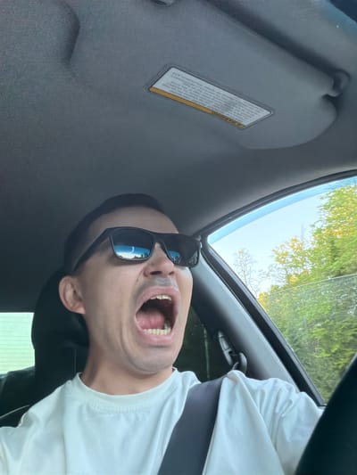 A man is sitting inside a car. He is wearing sunglasses and a white t-shirt. His mouth is open. The car is in an unknown location, with trees visible through the sunroof. The interior of the car is gray, with a sun visor on the left side. There is no information to indicate the location is in Severnyy, Russia.