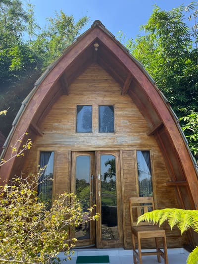 A wooden triangular-shaped cabin is set against a backdrop of lush green trees and a clear blue sky. The cabin features a large wooden door with glass panels and two smaller rectangular windows above it. To the right of the door, a simple wooden chair sits on a white tiled porch, partially obscured by a vibrant green fern. The cabin appears to be located in a tropical environment, possibly a resort or a homestay, given the surrounding vegetation and the outdoor seating. The overall scene suggests a tranquil setting in Tetebatu, Indonesia, a region known for its natural beauty and rice terraces.