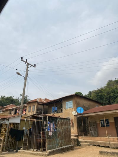 This image depicts a street scene in Okengwe - Obehira, Nigeria, under an overcast sky. The foreground features unpaved, light-colored ground.
In the mid-ground, a tall, grey concrete utility pole with a street light fixture stands prominently on the left, supporting multiple black power lines that stretch horizontally across the frame.
The central and right portions of the image are dominated by a cluster of single and two-story buildings, characterized by earth-toned, unpainted walls and rusted corrugated metal roofs.
* On the far left, a partial view of a building with a "ELECTRONICS SHOP" sign shows dark clothes hanging to dry on metal grates.
* Adjacent to this, a freestanding rectangular enclosure made of heavily rusted corrugated metal sheets with a barred metal gate is visible. Inside, clothes are hanging, and a dark object, possibly a sewing machine, is discernible.
* Behind this enclosure, a two-story building features faded, unpainted walls, a rusted corrugated roof, and a balcony or open area on the upper floor. Some windows have blue frames or reflective blue panes, while others appear boarded or shuttered. A white satellite dish is mounted on its facade.
* To the right, a one-story building with a reddish-brown corrugated metal roof has two-toned walls (lighter brown base, darker brown upper). It features windows with blue frames and security grates, and a dark door. A blue satellite dish with "GHS" branding is affixed to its exterior. Raised steps or a platform lead to this building.
Dense green trees are visible on an elevated terrain or hillside in the background, particularly on the right side of the frame. No individuals are visible within the scene.