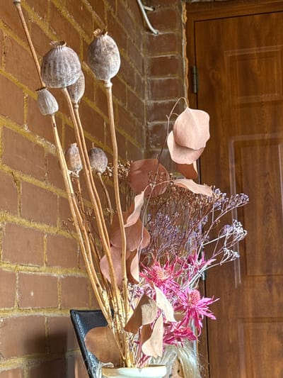 This image depicts a dried floral arrangement situated indoors in Anglininkai, Lithuania. The arrangement, centrally located, consists of several dried botanical elements. Prominently featured are multiple elongated, grayish-brown poppy seed pods on slender, dried stems of varying lengths. These are accompanied by several broad, light reddish-brown dried leaves, possibly from eucalyptus, and numerous delicate clusters of small, dried purple and white florets on thin, branching stems. Additionally, several vibrant magenta-pink thistle-like dried flowers with spiky petals are integrated into the composition. The base of the arrangement is held in a partially visible, light-colored vase at the bottom center.
The background on the left side of the frame features a reddish-brown brick wall with yellowish mortar lines. A white electrical cable runs vertically along the upper left portion of this wall. To the right, a medium-brown wooden door with a vertical paneled design is visible, along with a silver-colored hinge on its left frame. A strip of yellowish insulation foam is present in the gap between the brick wall and the door frame. A portion of a black, textured object, likely a chair back, is visible behind the vase on the lower left. Strands of blonde hair are faintly discernible at the very bottom right edge of the image. The scene is uniformly lit by ambient light, accentuating the textures of the dried plants and background surfaces.