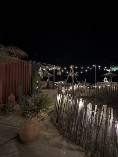 The image depicts an outdoor area at night, possibly a restaurant or bar, located in Torreilles, France. The scene is illuminated by strings of lights and a disco ball suspended from a wooden structure. A rough, wooden fence runs along the foreground, with several lights attached to it. Behind the fence, a sandy area is visible. To the left, a large terracotta pot holds a spiky plant, and a red shipping container with some wooden structures and cacti are present. Further in the background, there are more wooden structures, thatched umbrellas, and seating areas. The overall atmosphere is rustic and bohemian.
