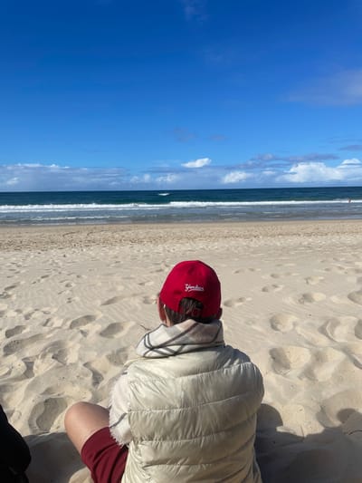 A person is sitting on a sandy beach in Surfers Paradise, Australia, facing the ocean. The person is wearing a red New York Yankees baseball cap, a light-colored puffer vest over a scarf, and maroon shorts. The sand is marked with footprints. The ocean is a deep blue with white waves breaking on the shore. The sky above is a clear blue with scattered white clouds. In the distance, a few people can be seen in the water. The overall impression is one of a relaxed day at the beach.