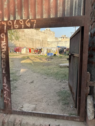 View through a rusty, metal frame, possibly a doorway or window, revealing an impoverished area in Mathura, India. The ground is dirt with sparse grass patches. Temporary shelters, constructed from various materials, line the edge of the open area, suggesting informal housing or a slum. A tall, unfinished concrete building forms the backdrop, with clotheslines and assorted discarded items visible. The scene suggests poverty and makeshift living conditions.
