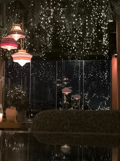The image captures a serene nighttime scene, likely from a restaurant or an event space in Palaio Faliro, Greece. The foreground features a dark, reflective surface, possibly a polished table or a water feature, reflecting the ambient lights. Above this, a dark, textured, hedge-like element spans horizontally across the frame.

The dominant features of the upper half of the image are numerous small, white fairy lights or string lights interwoven with green foliage, creating a dense, twinkling canopy overhead. Hanging from this canopy, to the left, are three distinctive, multi-colored pendant lamps. These lamps have a mid-century modern aesthetic, with layered shades in shades of pink, orange, and cream, emitting a soft, warm glow.

Behind the immediate interior space, large glass panels or windows reveal more of the outside, which is also illuminated by numerous small, distant lights, creating a sparkling backdrop. Through these windows, reflections and distant objects can be vaguely discerned, including more hanging light fixtures similar to those inside, suggesting an expansive decorated area. To the far left, a soft, warm light from what appears to be a candle or small lamp sits on a wooden surface, adding to the cozy atmosphere.

The time of day is clearly night or late evening, highlighted by the dark background and the prominence of artificial illumination. The overall ambiance is festive, warm, and inviting, suggesting a leisurely dining or social setting. No people or visible text are present in the image.