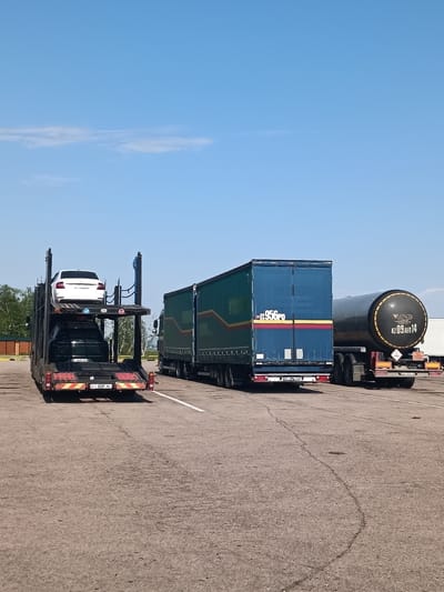 The scene captured is an outdoor parking area under a clear blue sky with a few wispy clouds. Three large vehicles occupy the space. From left to right:
1. A car carrier trailer laden with two cars. The upper car is white, while the lower one is dark.
2. A large green and blue semi-trailer truck, partially visible with a distinctive yellow and red stripe.
3. A black tanker truck with white markings, labeled "KZ 09 AED 14."
The parking surface appears to be asphalt with visible lines. There are trees and structures in the far background, indicating an area that may be an industrial park. The presence of license plates "KZ" implies the location to be Kazakhstan, specifically the Алтынды ауылдық округі, as requested.