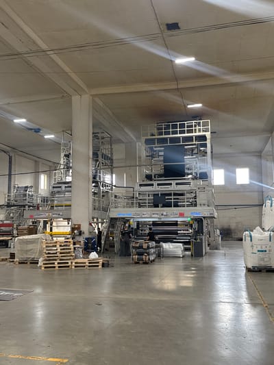 The image depicts the interior of a factory or manufacturing facility, likely a plastics extrusion plant, located in Osentsy, Russia. The vast space is characterized by a high concrete ceiling with exposed beams and fluorescent lighting, some of which appear to be newer LED fixtures. Various industrial machines, primarily tall, multi-story plastic film extrusion lines, dominate the scene. These machines are constructed with metal frames and appear to be in operation or ready for operation, with rolls of material visible.

Several individuals are present in the factory. Two men are seen working near one of the machines on the right, one appearing to be operating the equipment and the other standing nearby. Another individual is visible on the left side of the frame, also near a machine. The floor is a polished concrete surface, marked with yellow lines for traffic or safety.

In the foreground and to the left, stacks of pallets, some covered with plastic sheeting, are visible, suggesting incoming or outgoing raw materials or finished products. On the right side of the image, large white bulk bags, likely containing raw plastic pellets or similar materials, are stacked on pallets. The overall environment appears functional and industrial, with a focus on machinery and materials related to plastic production. The lighting casts reflections on the floor, highlighting the industrial nature of the setting.