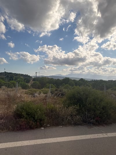 The image depicts a landscape scene in the Municipal Unit of Opountioi, Greece. In the foreground, a paved road with a white line is visible. Beyond the road, there is dry, tall grass and bushes. In the mid-ground, a densely vegetated area with various shades of green foliage stretches across the frame, interrupted by a wire fence with thin posts. Further back, rolling hills are covered with trees and dotted with wind turbines, some of which are clearly visible. In the distance, hazy mountains form the horizon, with a bright sky filled with scattered white and grey clouds above. The overall impression is of a rural or semi-rural area with natural and man-made elements. There are no people or animals present in the image.