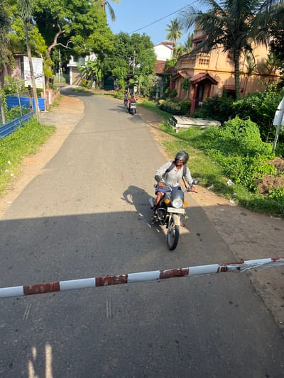 A man rides a motorcycle on a paved road in Kalutara, Sri Lanka. He is wearing a grey long-sleeved shirt, blue shorts, and a black helmet. The motorcycle is a silver and black model with a white license plate. The road is lined with lush green vegetation and trees, with houses visible in the background. The sky is clear and blue. A barrier with red and white stripes is visible in the foreground.