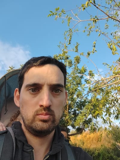 A man's selfie captures him from a low angle against an outdoor backdrop. He has dark hair, a beard, and light-colored eyes. He wears a dark jacket or shirt with visible straps or backpack elements. The background includes foliage with green leaves and golden grasses, suggesting a natural environment. A clear blue sky with some wispy clouds is visible above the trees. The photo has natural lighting, giving it a vibrant feel.