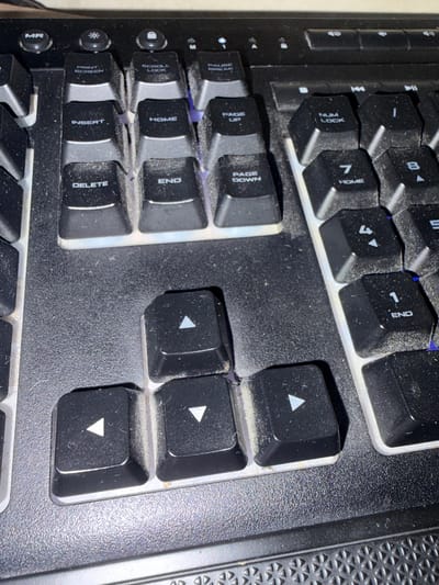The image shows a close-up of a black computer keyboard with several keys visible. The keys are dusty and some have illuminated backlighting. Specific keys visible include "MR", "PRINT SCREEN", "INSERT", "DELETE", "SCROLL LOCK", "HOME", "END", "PAGE UP", "PAGE DOWN", "NUM LOCK", and numerical keys. There are also arrow keys visible at the bottom of the image. The keyboard appears to be a standard QWERTY layout. The environment is dimly lit, suggesting indoor use. There is no indication of location in the city of Mirnoye, Ukraine, as this is a general photograph of a keyboard. There are no people or actions depicted.