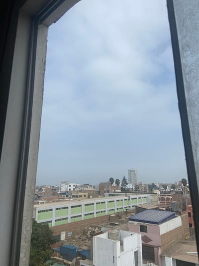 The image captures a view from inside a building, looking out through a window. The sky, which is predominantly overcast, fills a large portion of the frame. Below the sky, a densely populated urban landscape stretches into the distance. Buildings of varying heights and architectural styles crowd together, creating a complex cityscape. A notable building, distinguished by its multiple stories and alternating green and white façade, stands out. It is in the city of San Miguel, Peru. Other structures include residential buildings, some with visible roof details. At the edge of the view, a hint of greenery is present, suggesting the presence of trees. The window frame, visible at the top and bottom of the image, provides a border to the scene.