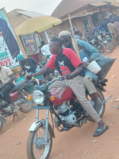 People sit on a red motorcycle in a dusty area. A person in a red and black shirt is near the front, facing forward. A man lies on top, with his legs stretched back. Other motorbikes surround them, with people nearby. Buildings and signs are visible in the background. The overall scene appears to be a casual gathering in a busy or market setting.