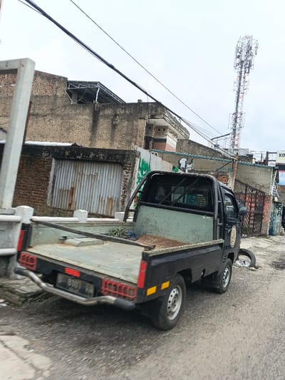A black pickup truck is parked on a paved road next to a curb. The truck bed appears to be metallic with a faded green interior. Behind the truck, there is a weathered building with corrugated metal and brick sections. Above the building, power lines crisscross the sky, leading towards a cellular tower in the distance. The scene is overcast.