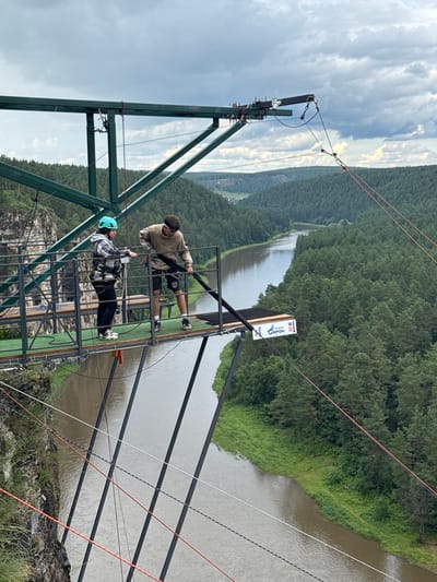 The image shows a bungee jumping platform on a metal structure overlooking a wide, tree-lined river in Kul'metovo, Russia. Two individuals are on the platform. One person, wearing a blue helmet and a checkered jacket with a safety harness, stands near the railing. The other person, wearing a light brown hoodie and dark shorts, is holding a thick black rope, appearing to prepare for a jump. The river below is flanked by dense green forest, and the sky is overcast with dramatic clouds. The platform is equipped with safety features like railings and ropes. A sign on the platform indicates "Grifon" and "80 M," suggesting it is a bungee jumping site with a significant drop.
