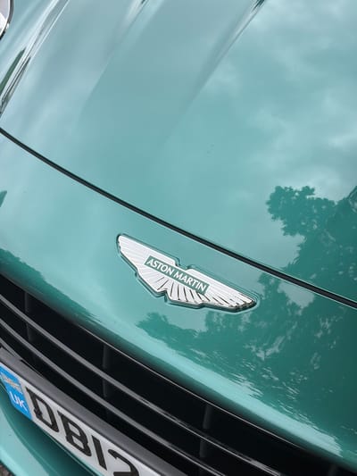 A close-up, angled shot captures the front detail of a teal or mint green Aston Martin car. The iconic Aston Martin winged badge is prominently featured in the upper middle of the frame, displaying "ASTON MARTIN" text in white letters against a dark background, bordered by chrome.

The car's highly reflective paint surface shows clear reflections of a cloudy sky and tree branches, suggesting an outdoor setting during daytime. A dark, slatted grill is visible to the right of the badge, extending horizontally across the lower right quadrant of the image. Partially visible below the grill is a white UK license plate, featuring the blue strip with the Union Jack flag and "UK" identifier. The visible characters of the license plate appear to be "DB17" or "DBI2". The image is taken from a low angle, emphasizing the sleek lines and shiny finish of the vehicle. The scene is static, focusing on the car's aesthetic details. The context provided indicates the location is Harrogate, United Kingdom.