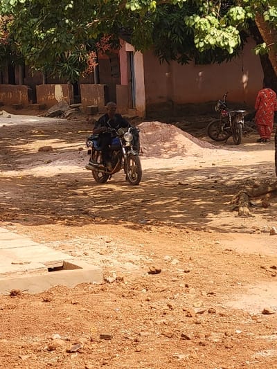 This image depicts an outdoor scene in Ebenebe, Nigeria, under bright daylight conditions, evident by strong shadows on the reddish-brown unpaved ground. A concrete drainage structure is visible in the lower-left foreground. The central subject is an adult male, appearing middle-aged to elderly, with dark skin and short dark hair, wearing a dark-colored top and dark trousers. He is seated on a blue-framed motorcycle with silver components, facing slightly right of the viewer. Behind him and to his right, a mound of light-colored sand or gravel is present. In the background, a single-story building with light brown walls and a pink door frame is partially visible, with green foliage from an overhead tree obscuring its upper portion. To the right of this building, a second, dark-colored motorcycle is parked. A person in a red patterned garment is seen standing near this parked motorcycle, facing away from the camera. Further to the left of the main building, partial concrete or mud-brick structures are observed, and tree roots are visible on the far right of the foreground.
