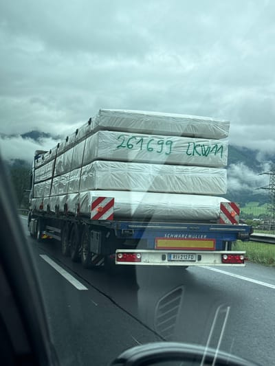 A semi-trailer truck is driving on a wet highway, likely in Kammern im Liesingtal, Austria, given the presence of mountains and electric pylons in the background, indicative of an alpine region. The truck is transporting a large, stacked load of rectangular items, possibly building materials or mattresses, wrapped in white plastic. Green markings on the top layer of the load read "261699 LKW". The truck itself has "SCHWARZMÜLLER" written on the rear, and a license plate reading "RI 212 FD". The sky is overcast and the road surface is dark and reflective, suggesting recent rain. The image is taken from another vehicle on the highway.