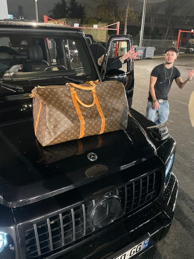 A black Mercedes-Benz G-Wagon is parked, with a large brown Louis Vuitton duffel bag placed on its hood. A man stands leaning on the car, wearing a black t-shirt, jeans, and jewelry. He is making a hand gesture towards the camera. Another person is visible in the driver's seat, with their hands raised in the air. The car is parked on a street with other vehicles and buildings in the background. The scene seems to be taken at night, lit by streetlights.