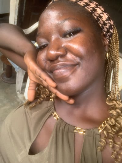 In Eliozu, Nigeria, a close-up, waist-up selfie captures a woman with dark skin and braided hair. She is wearing a khaki-colored garment with a gold chain detail at the neckline. Her left arm is bent, and her hand rests gently on her cheek, with her fingers slightly splayed. She is looking towards the camera with a subtle smile. The background is out of focus but appears to be an indoor setting with muted colors, possibly a wall and furniture. The lighting is soft, highlighting the sheen on her skin.