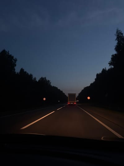 The image depicts a dark, rural highway at dusk or night, likely in Anatol'evka, Russia. The road is paved with visible white lane markings. On either side of the road, dense, dark forests line the highway, obscuring the background. In the distance, a large semi-truck with illuminated red taillights is driving away from the camera. Two circular traffic signs with red borders and white interiors are visible on the sides of the road, one on the left and one on the right, both appearing to be prohibitory signs. The sky above is a deep blue, transitioning to darker hues as it reaches the top of the frame, with subtle cloud formations visible. The scene is lit by the truck's taillights and the headlights of the vehicle from which the photo is taken, casting an orange glow on the road surface.