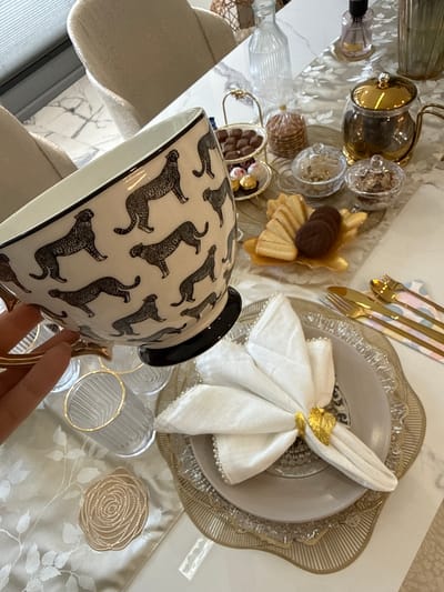 The image depicts a table setting with a white marble surface and a patterned table runner. Various decorative items are present, including glass containers with sweets, a plate with cookies and chocolates, and a napkin folded artistically on a decorative plate with gold-edged cutlery. A tea cup with a black and white leopard print design is prominently positioned in the foreground and held by a hand with polished nails. The background suggests an interior space with light-colored chairs. The scene is likely set indoors, possibly in a dining room or a retail display area. Nottingham, United Kingdom is not suggested by any details.