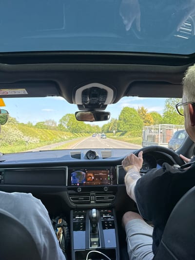 The image shows the view from inside a car driving on a road lined with green trees and bushes, under a blue sky in South Cambridgeshire, United Kingdom. The driver is holding the steering wheel. The car's dashboard features a central infotainment screen, air vents, and gear selector. A rearview mirror is visible. The driver is wearing glasses and a dark-colored shirt. A passenger is visible in the front passenger seat, wearing a light-colored shirt.