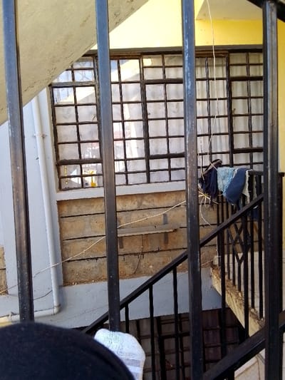 The scene depicts an architectural view from an elevated position within Kalimoni ward, Kenya, looking across what appears to be a residential building's exterior or an internal lightwell/stairwell. In the immediate foreground, multiple dark vertical metal bars of a railing are prominent, with the top of a dark-colored shoulder and a white sock visible at the bottom-center, indicating the observer's immediate proximity.

The midground features a large, rectangular window composed of numerous small panes, secured by a dark grid-like frame and partially obscured by translucent light-colored curtains. Below this window, a wall constructed from horizontal courses of light brown concrete blocks is visible, exhibiting visible mortar lines. A white PVC drainage pipe runs vertically along the left side of this concrete block wall. A thin, light-colored wire spans horizontally across the upper section of the concrete block wall. Attached to the concrete block wall is a narrow, horizontal fixture, appearing to be a shelf or bracket.

To the right, a black metal staircase railing descends towards the bottom-right of the frame. Several pieces of fabric, specifically dark blue and light blue articles of clothing, are draped over this staircase railing. A single light-colored string or rope hangs vertically in front of the window near the center-right. The wall section immediately above the window is painted yellow. No identifiable individuals are present within the depicted scene beyond the partial view of the observer.
